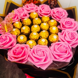 May include: A bouquet featuring pink roses and gold-wrapped chocolate candies. The arrangement includes a decorative butterfly and a gold "Happy Birthday" topper, all wrapped in black and gold paper with a pink ribbon.