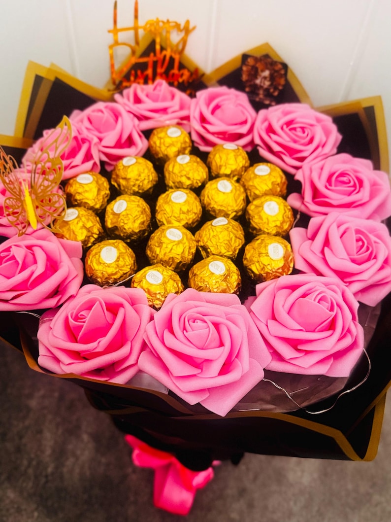 May include: A bouquet featuring pink roses and gold-wrapped chocolate candies. The arrangement includes a decorative butterfly and a gold "Happy Birthday" topper, all wrapped in black and gold paper with a pink ribbon.