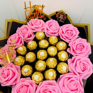 May include: A bouquet of pink roses and gold-wrapped chocolates. The arrangement includes a gold butterfly and a gold "Happy Birthday" topper. The bouquet is wrapped in black and gold paper.