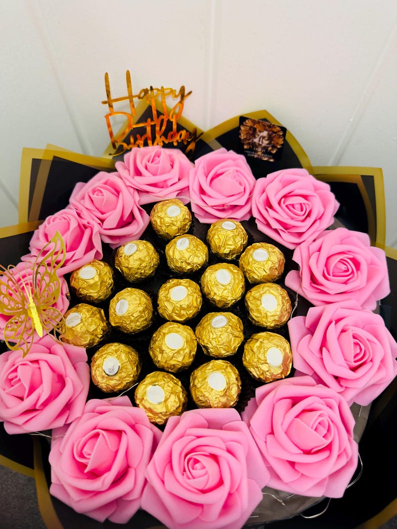 May include: A bouquet of pink roses and gold-wrapped chocolates. The arrangement includes a gold butterfly and a gold "Happy Birthday" topper. The bouquet is wrapped in black and gold paper.