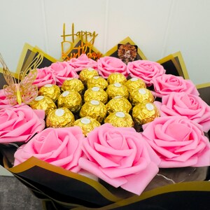 May include: A bouquet of pink roses and gold-wrapped chocolates. The arrangement includes a gold-colored butterfly and a gold-colored sign that says "Happy Birthday". The bouquet is wrapped in black and gold paper.