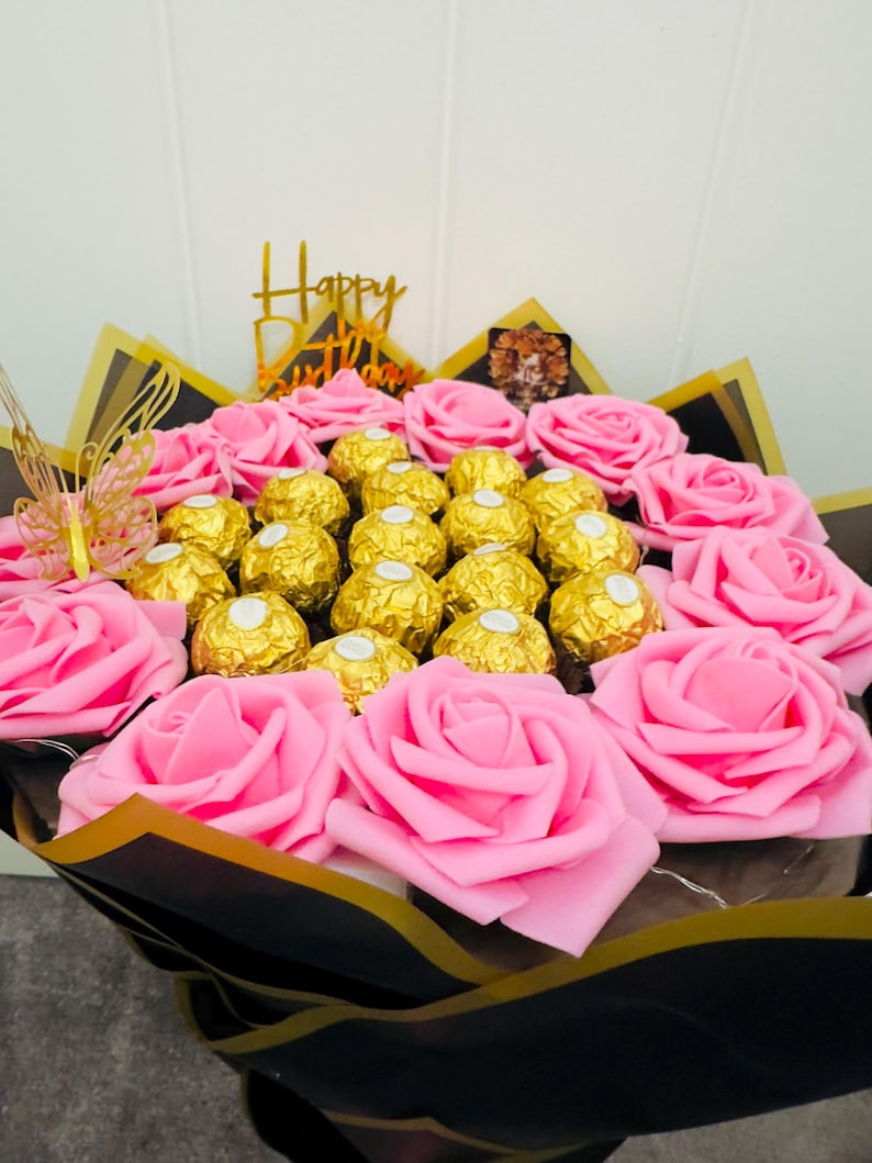 May include: A bouquet of pink roses and gold-wrapped chocolates. The arrangement includes a gold-colored butterfly and a gold-colored sign that says "Happy Birthday". The bouquet is wrapped in black and gold paper.