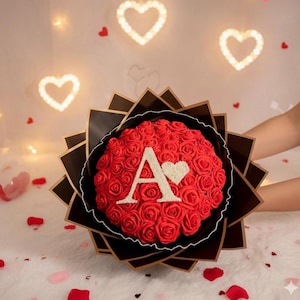 May include: A bouquet of red roses, circular in shape, with the letter "A" and a heart in the centre. The bouquet is wrapped in black and gold paper. The background features heart-shaped lights and scattered rose petals.