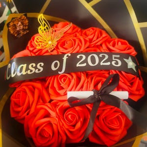 Pode incluir: Um buquê de rosas vermelhas com uma fita preta que diz "Class of 2025". O buquê é decorado com uma borboleta dourada e um pergaminho enrolado amarrado com uma fita marrom. O buquê é embrulhado em papel preto e dourado.