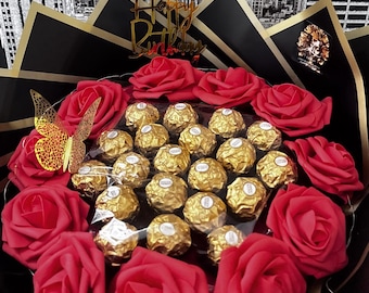 May include: A bouquet featuring red roses and gold-wrapped chocolates. The arrangement includes a gold butterfly accent and a gold "Happy Birthday" topper, all presented in black and gold wrapping.