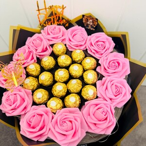 May include: A bouquet of pink roses and gold-wrapped chocolates. The arrangement includes a gold "Happy Birthday" topper and a decorative butterfly. The bouquet is wrapped in black and gold paper.