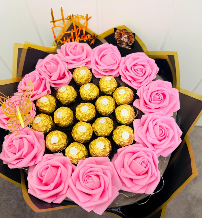 May include: A bouquet of pink roses and gold-wrapped chocolates. The arrangement includes a gold "Happy Birthday" topper and a decorative butterfly. The bouquet is wrapped in black and gold paper.