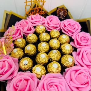 May include: A festive bouquet featuring pink roses and gold-wrapped chocolates. A gold-colored 'Happy Birthday' topper and a gold butterfly accent the arrangement. The bouquet is wrapped in black and gold paper.
