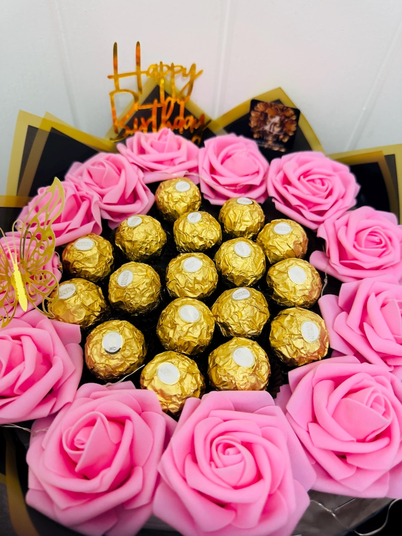May include: A festive bouquet featuring pink roses and gold-wrapped chocolates. A gold-colored 'Happy Birthday' topper and a gold butterfly accent the arrangement. The bouquet is wrapped in black and gold paper.