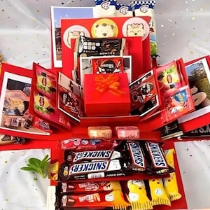 Może przedstawiać: Czerwone pudełko eksplodujące wypełnione różnymi smakołykami i prezentami. Pudełko zawiera batony Snickers, torebki herbaty, małe czerwone pudełko prezentowe z pomarańczową kokardą oraz inne różnorodne słodycze i przekąski. Elementy dekoracyjne obejmują naklejki z postaciami z kreskówek i małe świece.