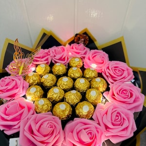 May include: A bouquet featuring pink roses and gold-wrapped chocolate candies. The arrangement includes a decorative gold butterfly and is wrapped in black and gold paper. Small lights are interspersed throughout the bouquet.