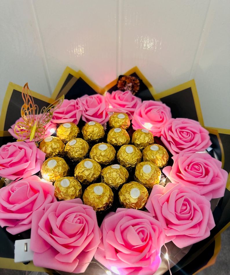 May include: A bouquet featuring pink roses and gold-wrapped chocolate candies. The arrangement includes a decorative gold butterfly and is wrapped in black and gold paper. Small lights are interspersed throughout the bouquet.