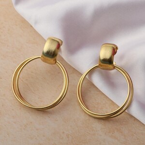May include: A pair of gold hoop earrings. Each earring has a rectangular top piece attached to a double-layered circular hoop. The earrings are presented on a light-coloured surface with white fabric in the background.