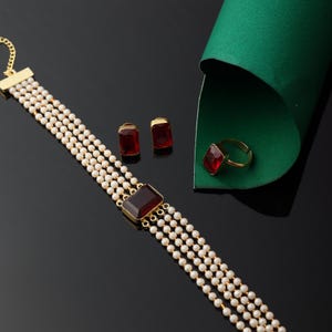 Op de afbeelding: Een goudkleurige sieradenset met een meerstrengs parelketting met een grote, rechthoekige, rode edelsteen. De set bevat bijpassende oorbellen en een ring, allemaal met rode edelstenen. De sieraden worden gepresenteerd op een zwart oppervlak.