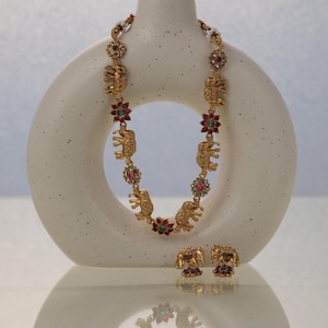 May include: Gold-coloured elephant necklace and matching earrings. The necklace features elephant pendants and floral accents with red, green, and clear gemstones. The earrings mirror the elephant and floral design. The jewellery is displayed on a white, abstract form.
