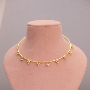 Puede incluir: Un collar gargantilla dorado con pequeñas perlas blancas colgantes. El collar se exhibe sobre un busto de maniquí rosa sobre un fondo neutro. El collar es de diseño sencillo y elegante.