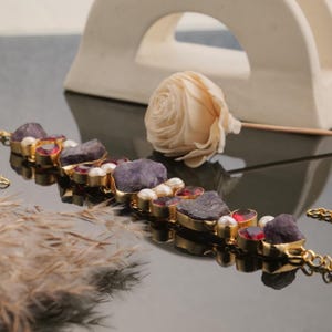 Könnte beinhalten: Ein goldfarbenes Armband mit einer Reihe von Edelsteinen. Das Armband enthält grob geschnittene, dunkelviolette Amethyststeine, kleine rote Edelsteine und weiße Perlen. Die Steine sind in goldenen Fassungen gefasst und schaffen ein luxuriöses und elegantes Design.