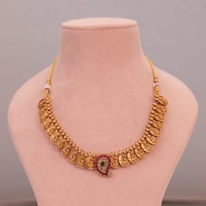May include: A gold-toned necklace featuring a series of coin-like pendants. The necklace has a central paisley-shaped pendant with red and green gemstones. The necklace is displayed on a pink mannequin.