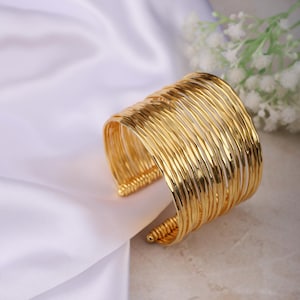 May include: A gold-coloured cuff bracelet. The bracelet is made of multiple parallel, textured metal bands. The ends of the bracelet have a decorative, coiled design. The bracelet is displayed on a white, silky fabric.