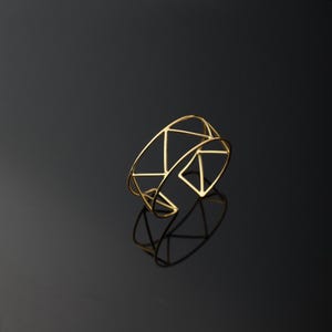 Könnte beinhalten: Ein goldfarbenes Armreif-Armband mit geometrischem Design. Der offene Armreif hat eine Reihe von miteinander verbundenen Dreiecken und Linien, die ein offenes, luftiges Design schaffen. Das Armband wird auf einer reflektierenden schwarzen Oberfläche präsentiert.
