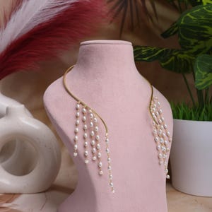 Op de afbeelding: Een goudkleurige ketting met watervalachtige witte parels, tentoongesteld op een roze mannequin. De ketting heeft een gebogen ontwerp dat om de nek drapeert, met meerdere strengen parels die naar beneden hangen. De achtergrond bevat planten en decoratieve elementen.