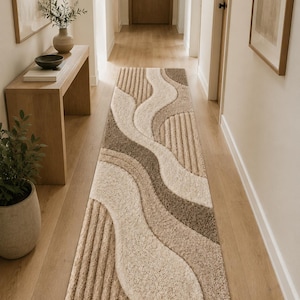 May include: A long rectangular runner rug featuring a textured, abstract wave pattern in shades of cream, beige, and light brown. The design incorporates alternating sections of high-pile tufted surfaces and linear, ribbed, or grooved textures.
