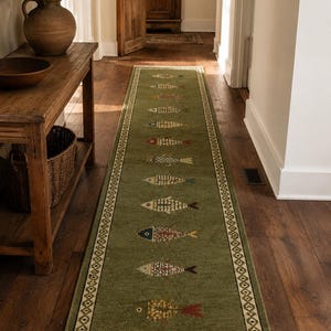 May include: A long, rectangular runner rug in olive green featuring a repeating pattern of stylized fish in various colors. The rug has a decorative geometric border and white fringe at both ends, laid flat on a dark wooden floor surface.