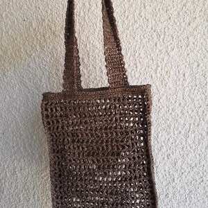 以下が含まれることがあります： 茶色のオープン織りのトートバッグ。2つの長いハンドルが付いています。白いテクスチャード加工の壁を背景にバッグが持たれています。バッグの織り方は網のような外観を作り出し、ハンドルはバッグと同じ色です。