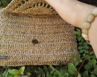 Könnte beinhalten: Goldfarbene, gehäkelte Clutch-Tasche mit Klappenverschluss. Die Tasche hat ein strukturiertes, gewebtes Aussehen und einen kleinen, runden Knopfverschluss. Die obere Klappe hat ein offenes, dekoratives Muster. Die Tasche wird vor grünem Laub gehalten.