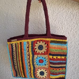 Può includere: Una borsa tote all'uncinetto fatta a mano con manico e finiture bordeaux. La borsa presenta un colorato design patchwork con quadrati e strisce nei toni del blu, giallo, arancione e marrone. La borsa è tenuta da una mano.