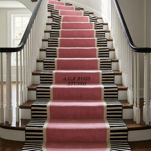 Peut inclure: Un tapis d'escalier sur mesure avec un panneau central rose uni bordé de rayures horizontales noires et blanches. Le tapis s'étend de la marche inférieure au sommet de l'escalier, avec le texte A.G.R RUGS STUDIO imprimé sur une marche.