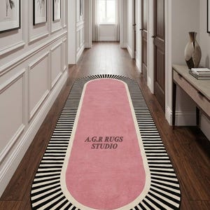 May include: An elongated oval runner rug featuring a solid pink center bordered by a thick cream band and an outer edge of radiating black and white stripes. The text A.G.R RUGS STUDIO is printed in dark gray in the center of the pink section.