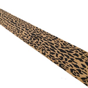 May include: A long, rectangular table runner with a tan background and black, irregular spots. The runner is made of a textured material and is angled diagonally across the frame. The overall design resembles a leopard print.