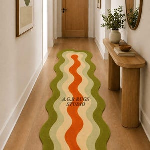 May include: A long, wavy runner rug featuring concentric, organic shapes in shades of dark green, light green, cream, and a central vibrant orange stripe. The text A.G.R RUGS STUDIO is printed in dark letters on the surface of the rug.