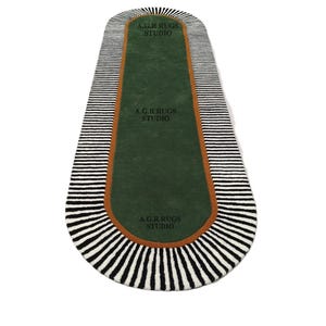 May include: An oval-shaped runner rug featuring a solid dark green center, a thin burnt orange border, and a wide outer edge with black and white radial stripes. The text A.G.R RUGS STUDIO is printed in black three times along the green surface.
