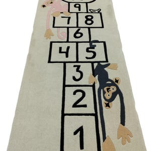 May include: A rectangular beige rug featuring a hopscotch design with black outlined boxes and numbers. Two cartoon monkeys are incorporated into the design, one pink and one dark gray. The rug has the text "A.O.R RUGS STUDIO".