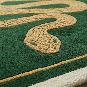 May include: A tufted rug featuring a deep green background with a large, winding tan snake design. The rug includes a tan border line and a thick white edge. Text on the rug reads A.G.R RUGS STUDIO, and a small white box in the corner displays Museum 100000305.
