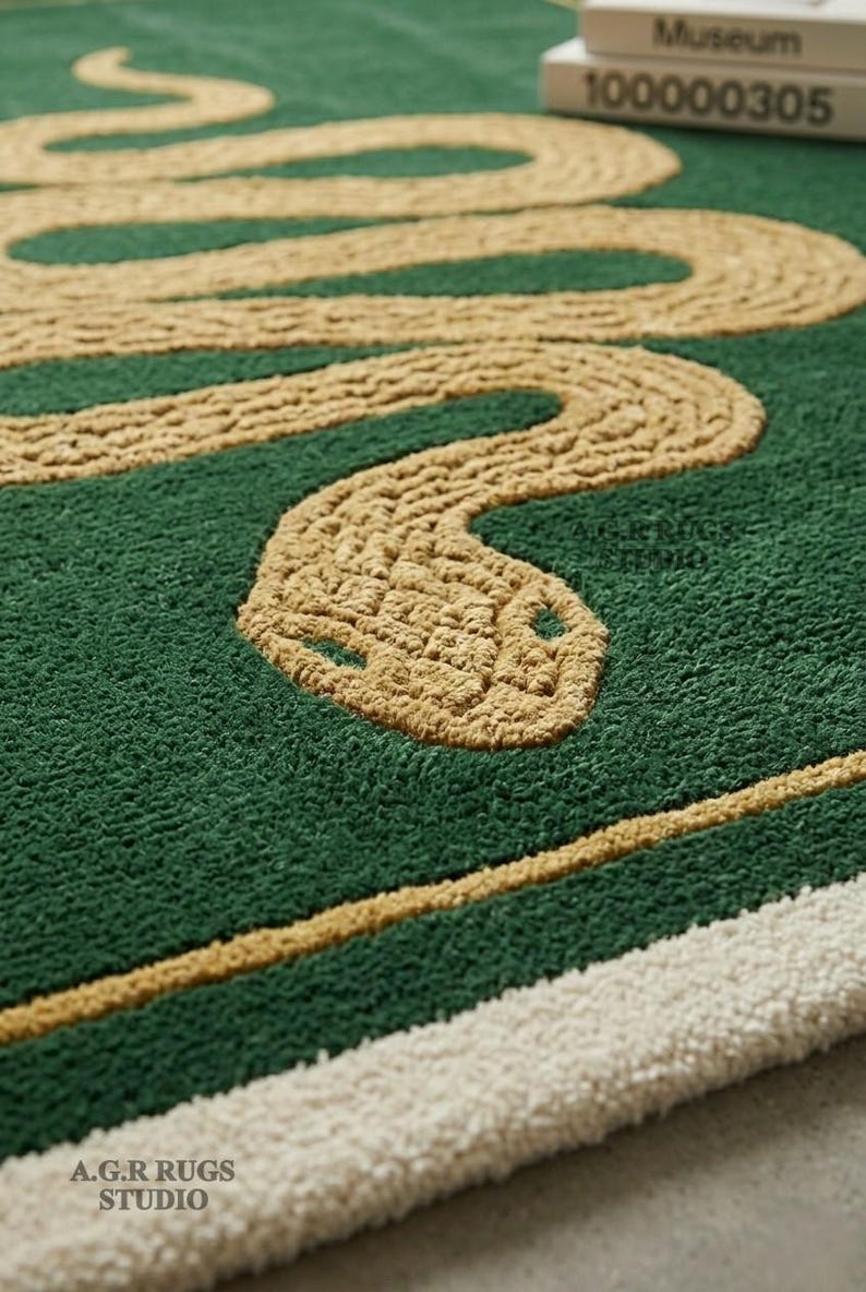 May include: A tufted rug featuring a deep green background with a large, winding tan snake design. The rug includes a tan border line and a thick white edge. Text on the rug reads A.G.R RUGS STUDIO, and a small white box in the corner displays Museum 100000305.