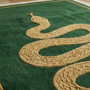 May include: A rectangular green rug featuring a textured, golden-tan coiled snake design in the center. The rug has a thin golden-tan border and the text A.G.R RUGS STUDIO is printed in black on the upper left side of the green surface.