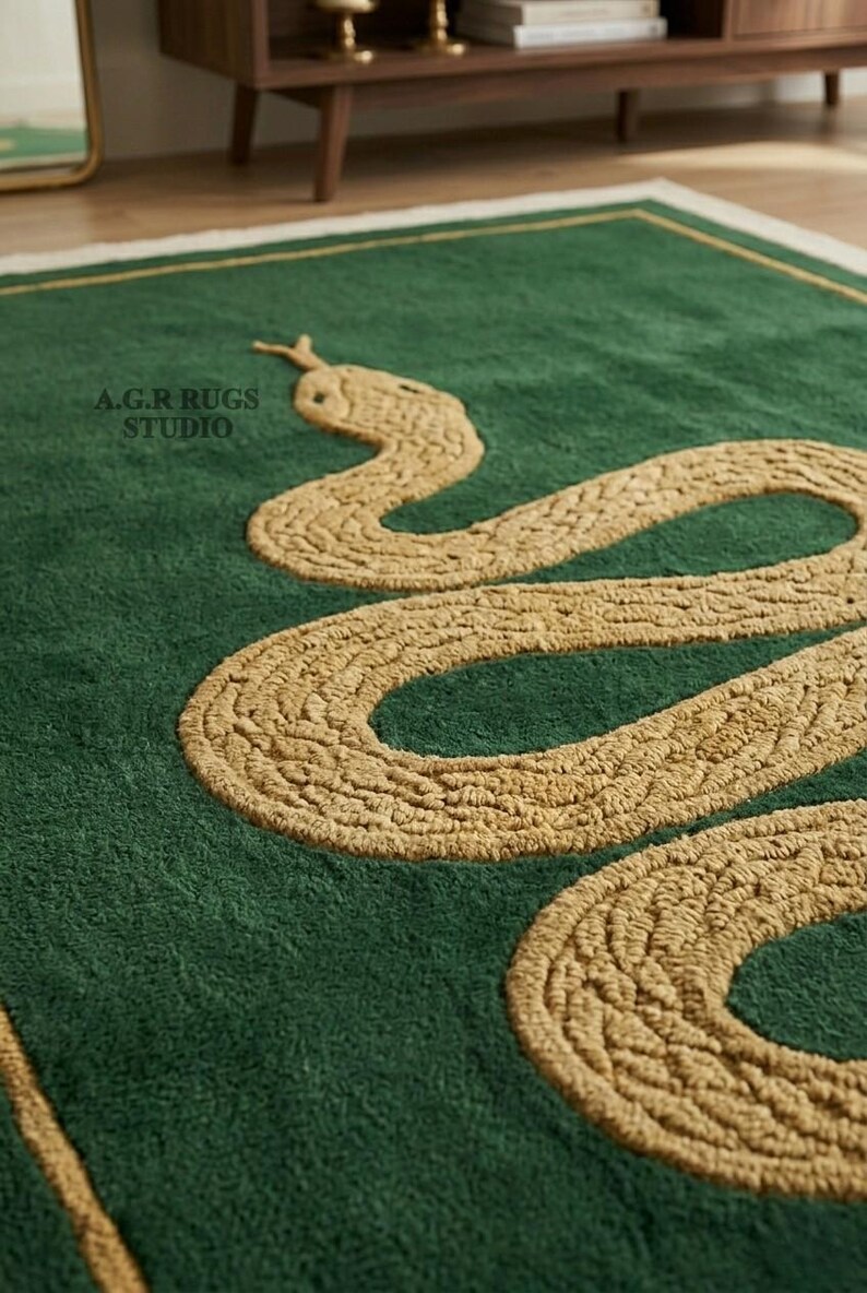 May include: A rectangular green rug featuring a textured, golden-tan coiled snake design in the center. The rug has a thin golden-tan border and the text A.G.R RUGS STUDIO is printed in black on the upper left side of the green surface.