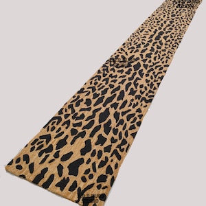 May include: A long, rectangular table runner with a tan background and a black leopard print pattern. The runner is made of a textured fabric and is suitable for home decor. The design is repeated across the entire length of the runner.