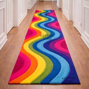 May include: A vibrant, multi-colored runner rug with a wavy design. The rug features a spectrum of colors, including pink, orange, yellow, green, blue, and purple. The text "A.G.R RUGS STUDIO" is visible on the rug.