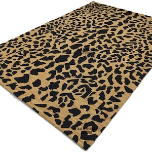 Può includere: Un tappeto rettangolare con sfondo beige e motivo leopardato nero. Il tappeto è angolato, mostrando la sua consistenza e il suo design. Le parole "A & R Studio" sono stampate sul tappeto.