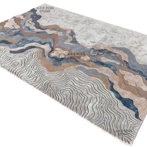 May include: A rectangular area rug with a landscape design. The rug features a pattern of flowing lines and shapes in shades of gray, blue, and brown. The text "A.G.R RUGS STUDIO" is visible in the image.