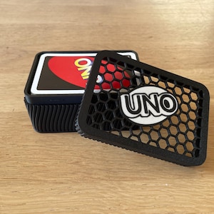 May include: Black UNO card game holder with a honeycomb lid design. The lid is partially open, revealing the UNO logo and a stack of cards. The holder is on a wooden surface.