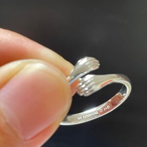 Puede incluir: Un anillo plateado con un dise&ntilde;o de dos manos abraz&aacute;ndose. El anillo est&aacute; sostenido por los dedos de una persona. El texto "Mi Amorcito J+D" est&aacute; grabado en el anillo.