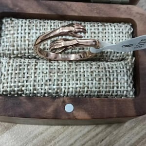 Puede incluir: Un anillo de oro rosa con un dise&ntilde;o de dos manos entrelazadas. El anillo, talla 8, se presenta en una caja de madera con un forro de tela tejida. El anillo est&aacute; grabado con las palabras "Always with you".