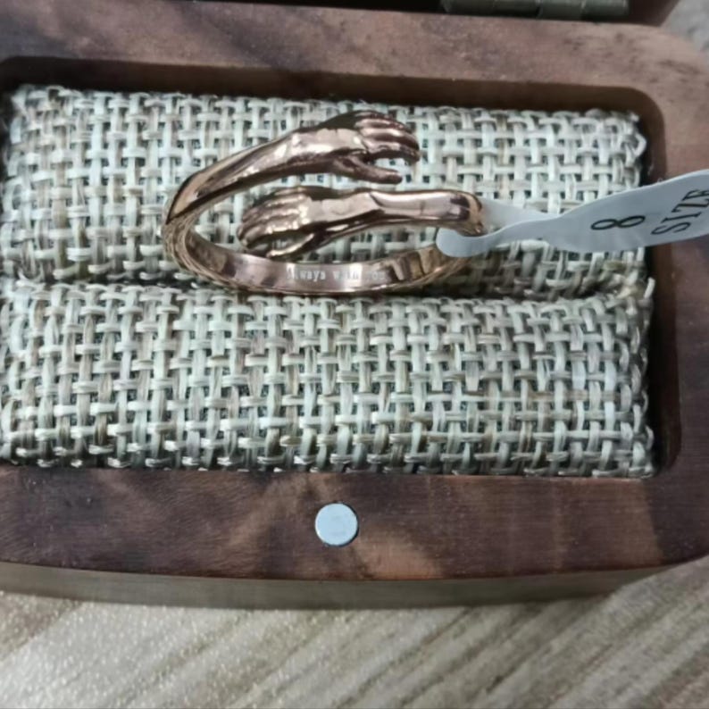 Puede incluir: Un anillo de oro rosa con un dise&ntilde;o de dos manos entrelazadas. El anillo, talla 8, se presenta en una caja de madera con un forro de tela tejida. El anillo est&aacute; grabado con las palabras "Always with you".