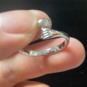 Puede incluir: Anillo plateado con un dise&ntilde;o de manos abraz&aacute;ndose. El anillo tiene la inscripci&oacute;n "Always With you x". El anillo est&aacute; sostenido por los dedos de una persona sobre un fondo oscuro.