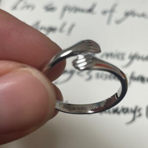 Puede incluir: Anillo plateado con forma de dos manos abraz&aacute;ndose. El anillo tiene grabadas las palabras "Always with you" en la banda, junto con un peque&ntilde;o s&iacute;mbolo de coraz&oacute;n. El anillo est&aacute; sostenido en la mano de una persona.